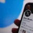 Elon Musk's twitter profile shown on a phone with the old Twitter bird in the background
